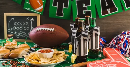 A Super Bowl party food spread featuring a football, beer bottles in referee jerseys, nachos, sliders, and football-themed decorations.