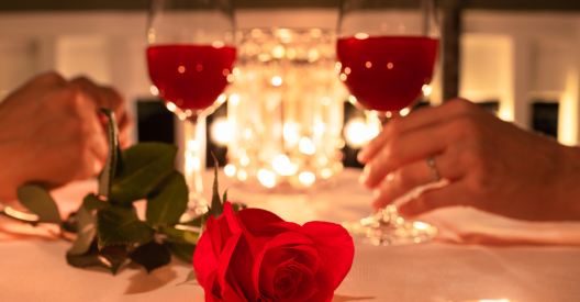 A romantic Valentine's Day dinner setting focusing on a red rose on a table, with a couple holding wine glasses in the soft-focus background.