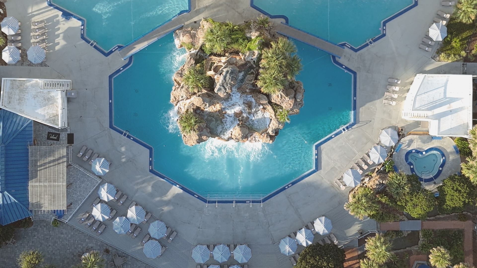 Top-down aerial drone view of a resort swimming pool featuring a central rock waterfall island and geometric pool edges surrounded by lounge chairs.