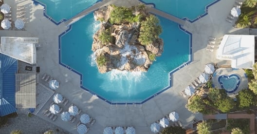 Top-down aerial drone view of a resort swimming pool featuring a central rock waterfall island and geometric pool edges surrounded by lounge chairs.