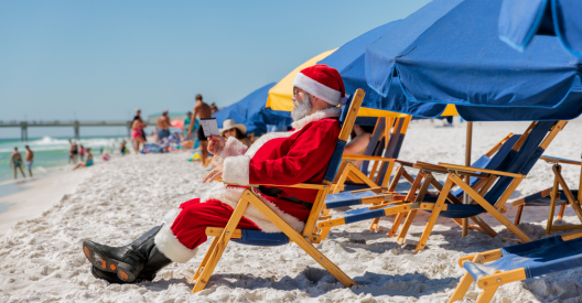Santa Claus relaxing on a wooden beach chair on white sand, reading a card while surrounded by blue and yellow beach umbrellas.