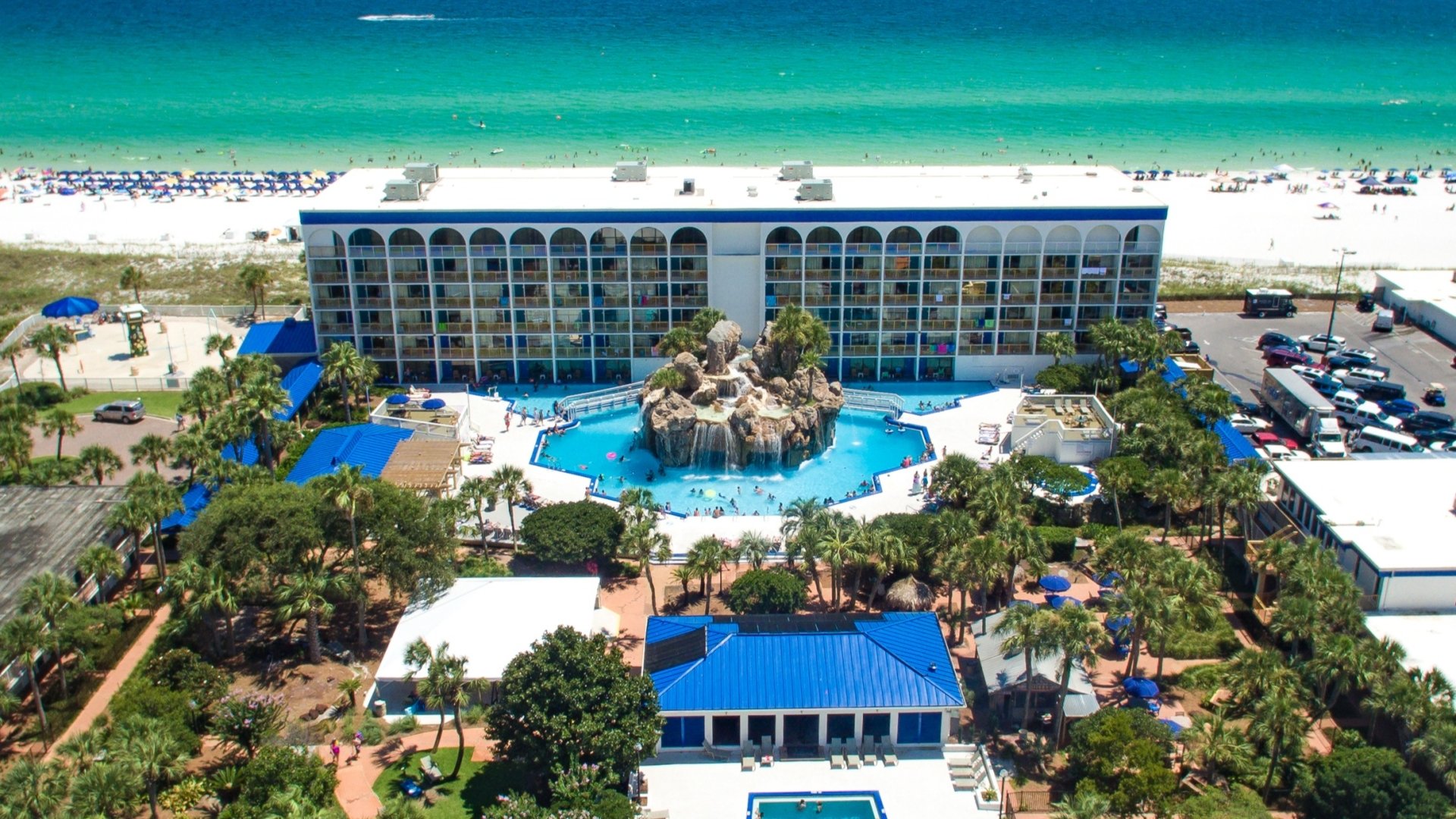Aerial drone view of a beachfront resort featuring a large swimming pool, a rock waterfall grotto, and a multi-story hotel building facing the ocean.
