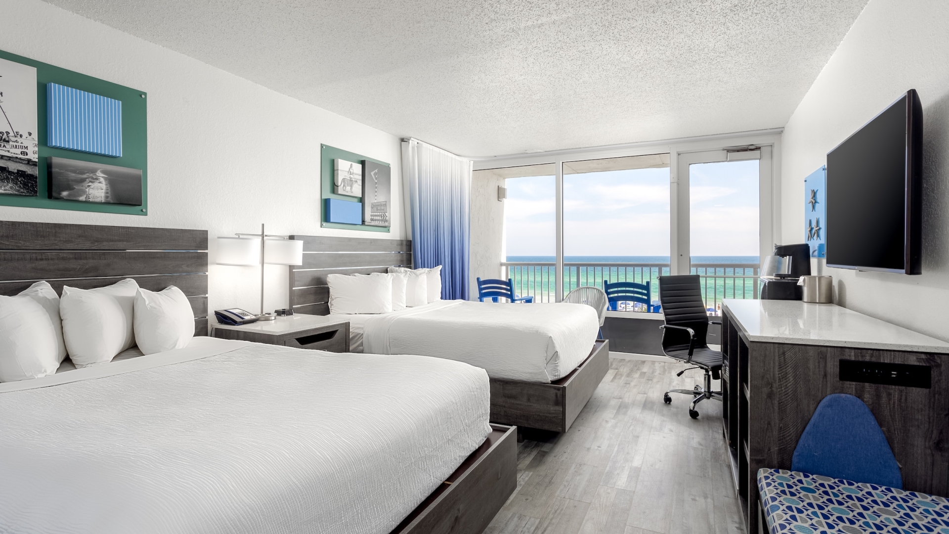 Modern double-queen hotel room with grey wood furniture, blue ombre curtains, and a sliding glass door leading to a balcony with an ocean view.