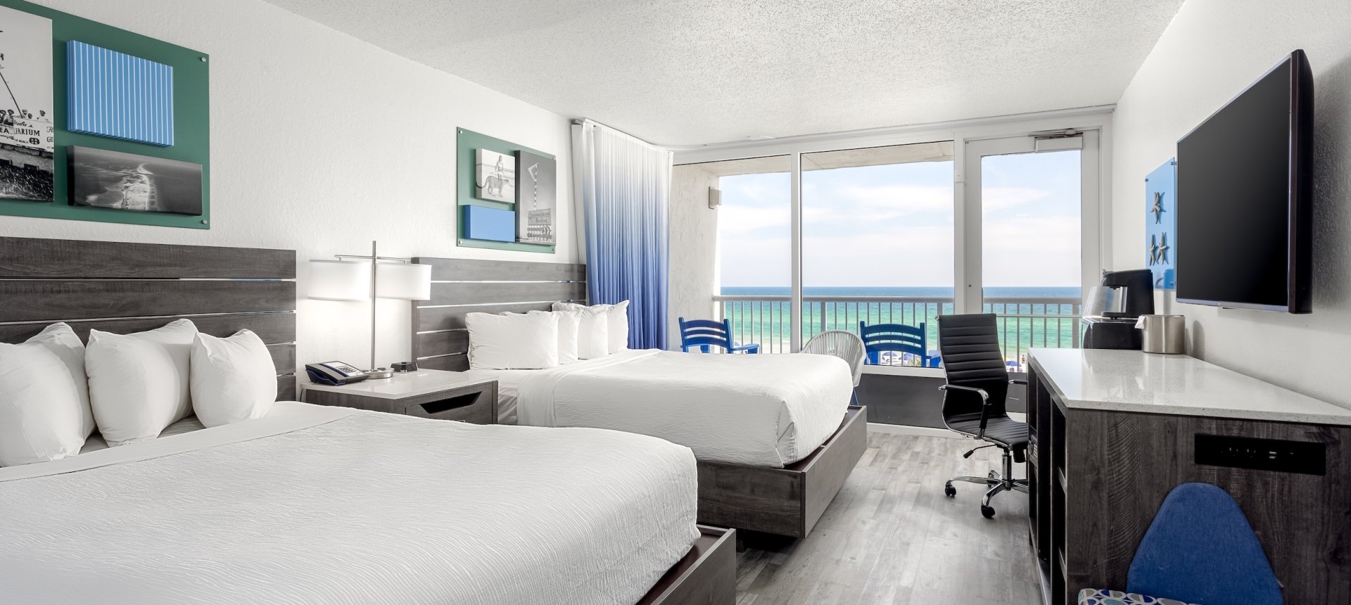 Modern double-queen hotel room with grey wood furniture, blue ombre curtains, and a sliding glass door leading to a balcony with an ocean view.