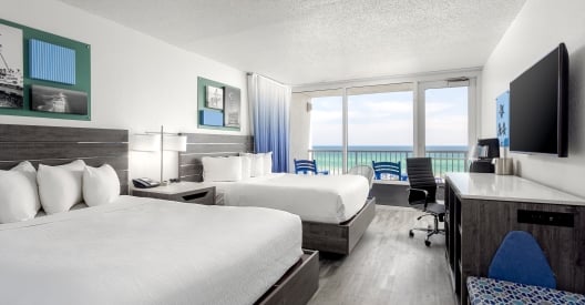 Modern double-queen hotel room with grey wood furniture, blue ombre curtains, and a sliding glass door leading to a balcony with an ocean view.