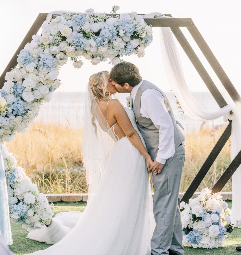 A romantic moment of a bride and groom kissing under a hexagonal wooden wedding arch decorated with lush white and light blue hydrangeas.