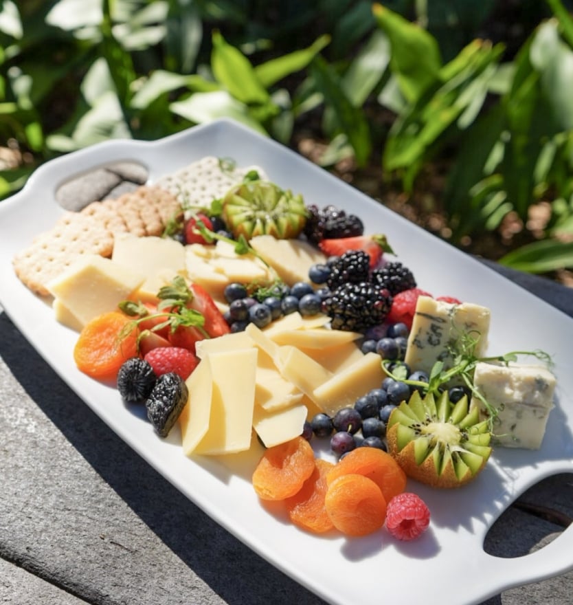 A colorful cheese board platter arranged with sliced cheeses, crackers, fresh berries, dried apricots, and kiwi garnishes on a white tray.