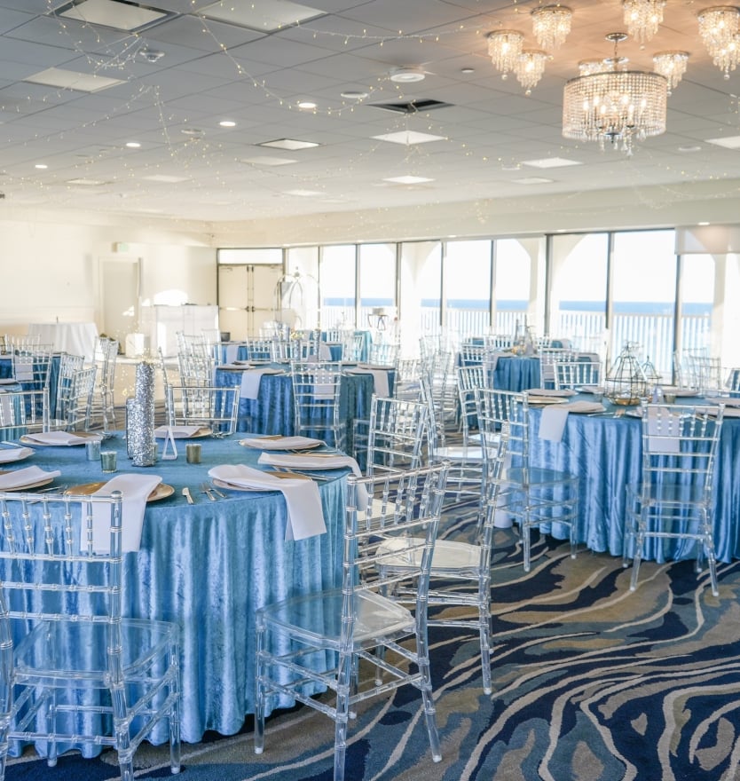 Wide angle view of a spacious hotel ballroom set for a wedding with blue linens, clear ghost chairs, ceiling string lights, and panoramic windows.