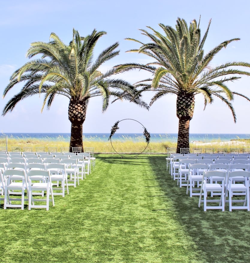 Outdoor beachside wedding ceremony setup featuring rows of white folding chairs on a green lawn, a circular metal arch, and two large palm trees framing an ocean view.