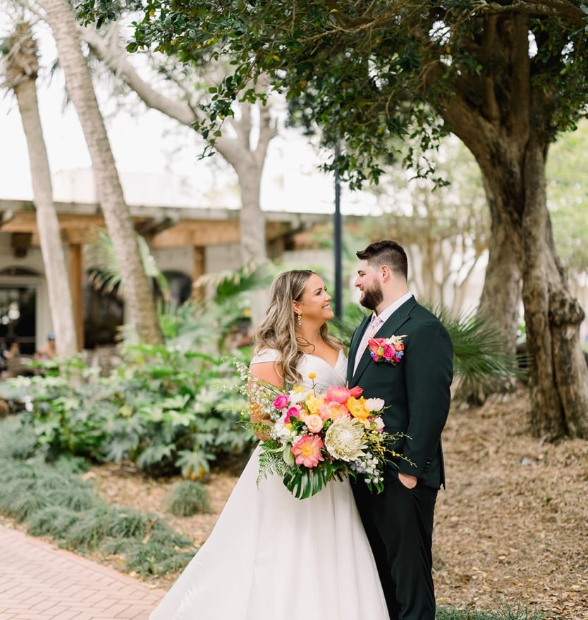 A bride and groom in wedding attire running barefoot and laughing along a white sandy beach with ocean waves in the background.