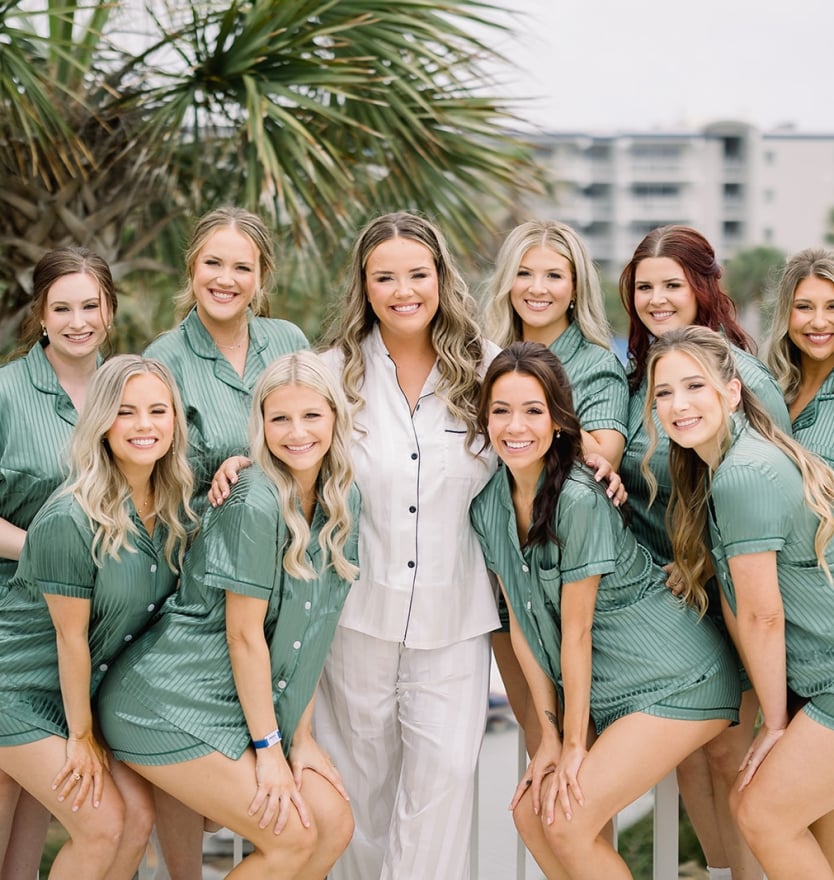 A smiling bride in white pajamas posing on a balcony with her bridesmaids, who are wearing matching sage green silky pajama sets.