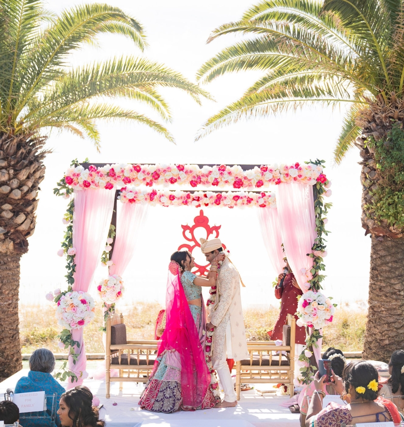 A bride and groom standing together under a vibrant pink and floral Mandap during a traditional Indian wedding ceremony, framed by two large palm trees.