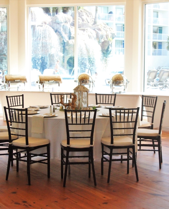 Bright indoor reception space featuring wooden flooring, round tables with black Chiavari chairs, and large windows overlooking a rock waterfall pool feature.