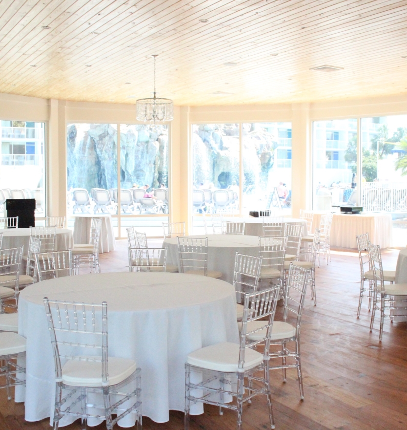Bright indoor event space with light wooden plank ceilings, white round tables with clear chairs, and large windows overlooking a rock waterfall feature.