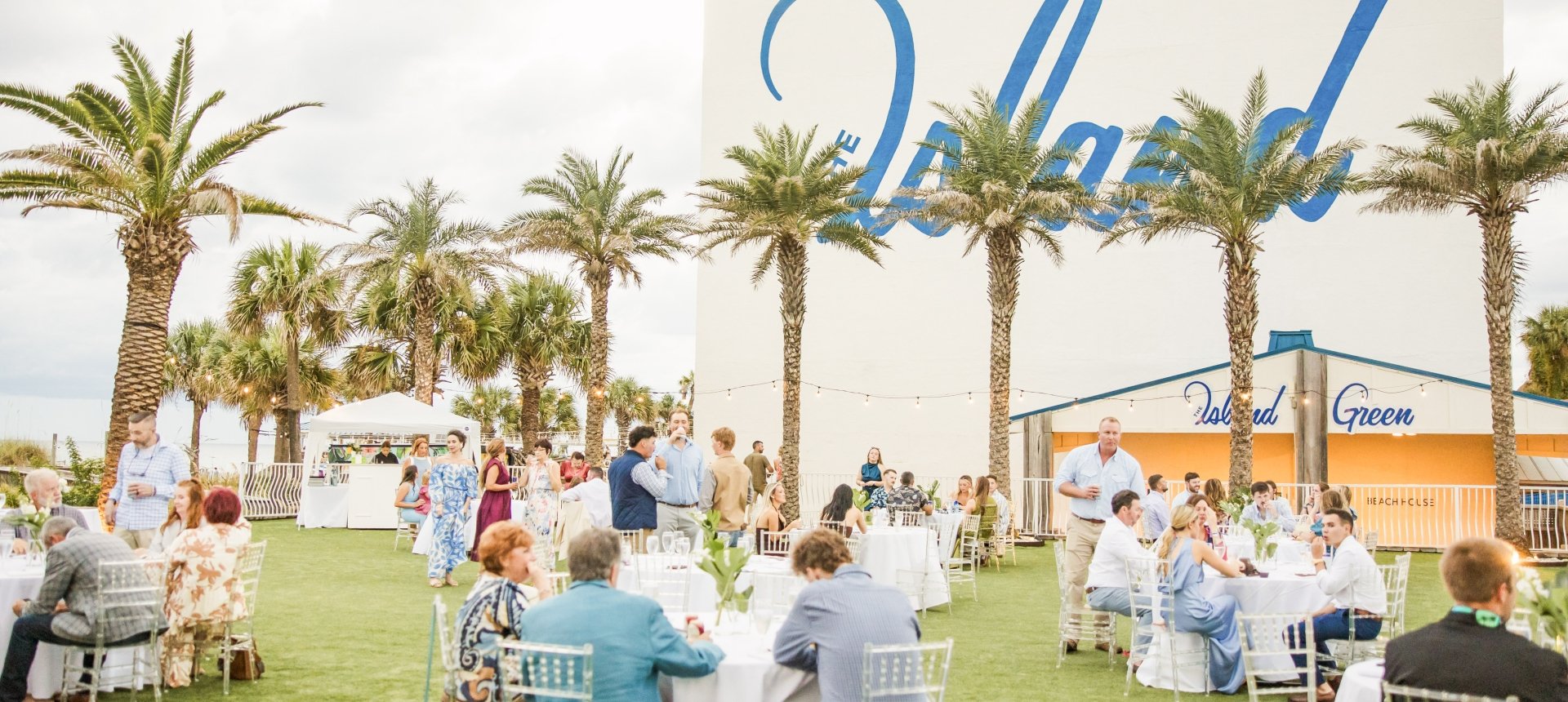 Outdoor wedding reception on a grassy lawn with guests seated at round tables, surrounded by palm trees and a large white building with blue 