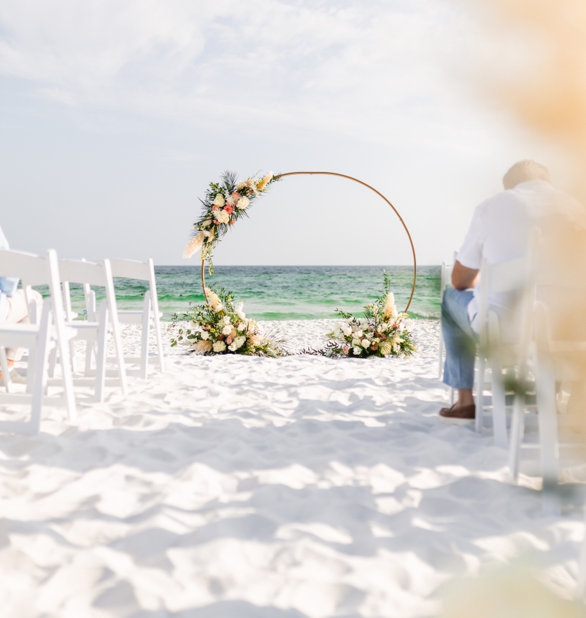 Scenic beach wedding ceremony setup on white sand, featuring rows of white folding chairs and a circular gold arch adorned with flowers facing the ocean.