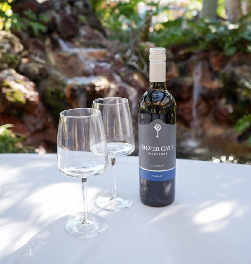 A bottle of Silver Gate Merlot wine standing next to two empty wine glasses on a white table, with a blurred rock waterfall in the background.